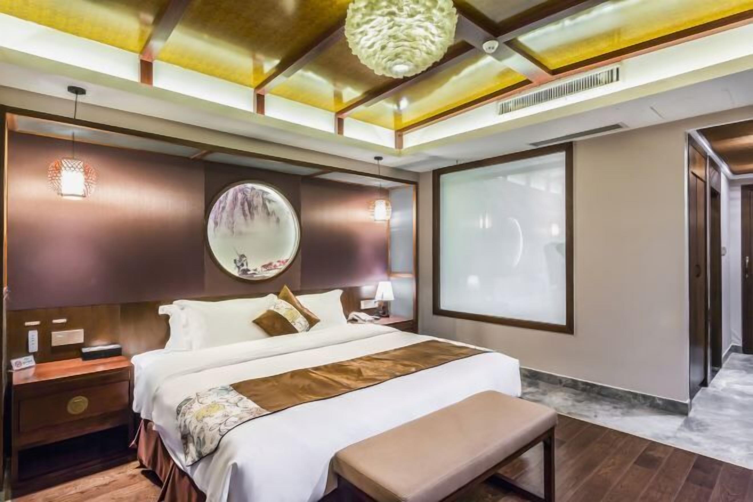 A modern hotel room with a large bed, a round mirror, a bench, and a large window.