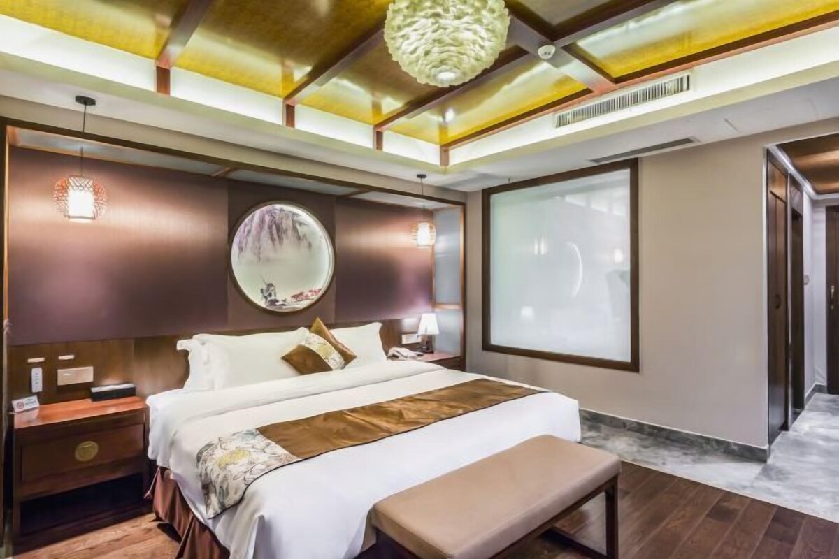 A modern hotel room with a large bed, a round mirror, a bench, and a large window.