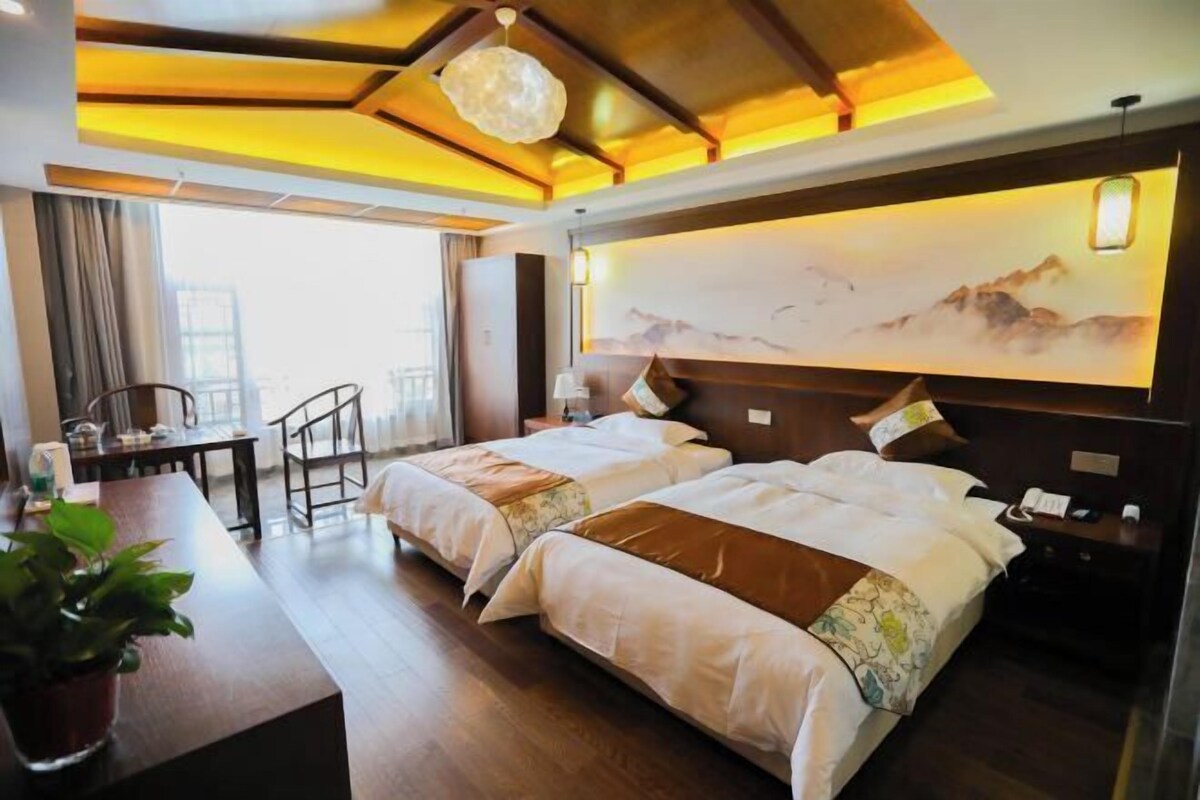 A hotel room with two beds, a desk with a chair, a large window, and a painting on the wall.