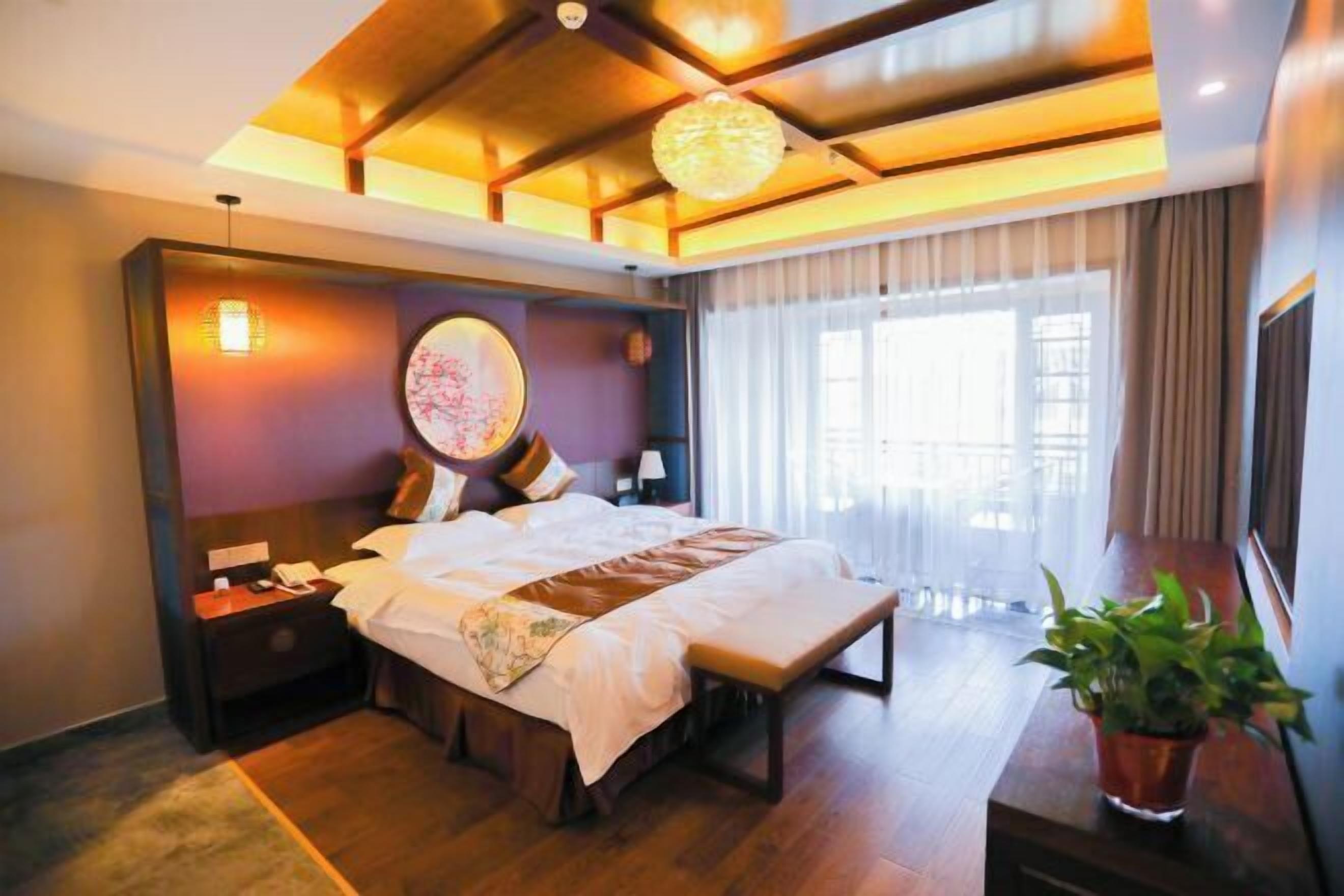 A hotel room with a large bed, bedside tables, a bedside lamp, a window with curtains, and a potted plant.