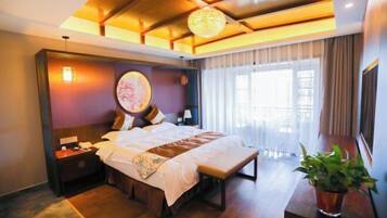A hotel room with a large bed, bedside tables, a bedside lamp, a window with curtains, and a potted plant.