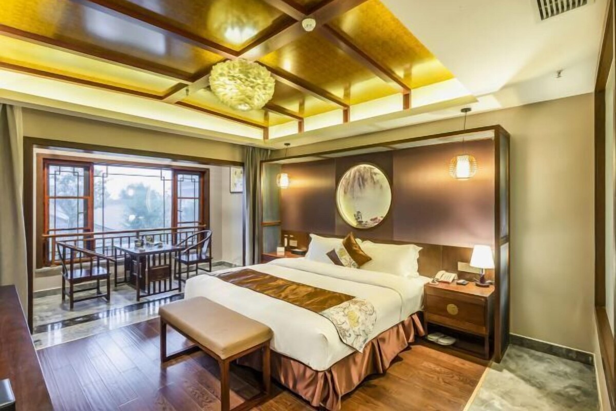 A hotel room with a large bed, a bedside table, a desk with chairs, and a view of the outdoors through large windows.