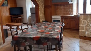 Un coin repas avec une table recouverte d’une nappe à motifs, entourée de chaises en bois. On y trouve un mur en pierre, un espace cuisine avec un fourneau et des placards, ainsi qu’un téléviseur posé sur un meuble en bois.