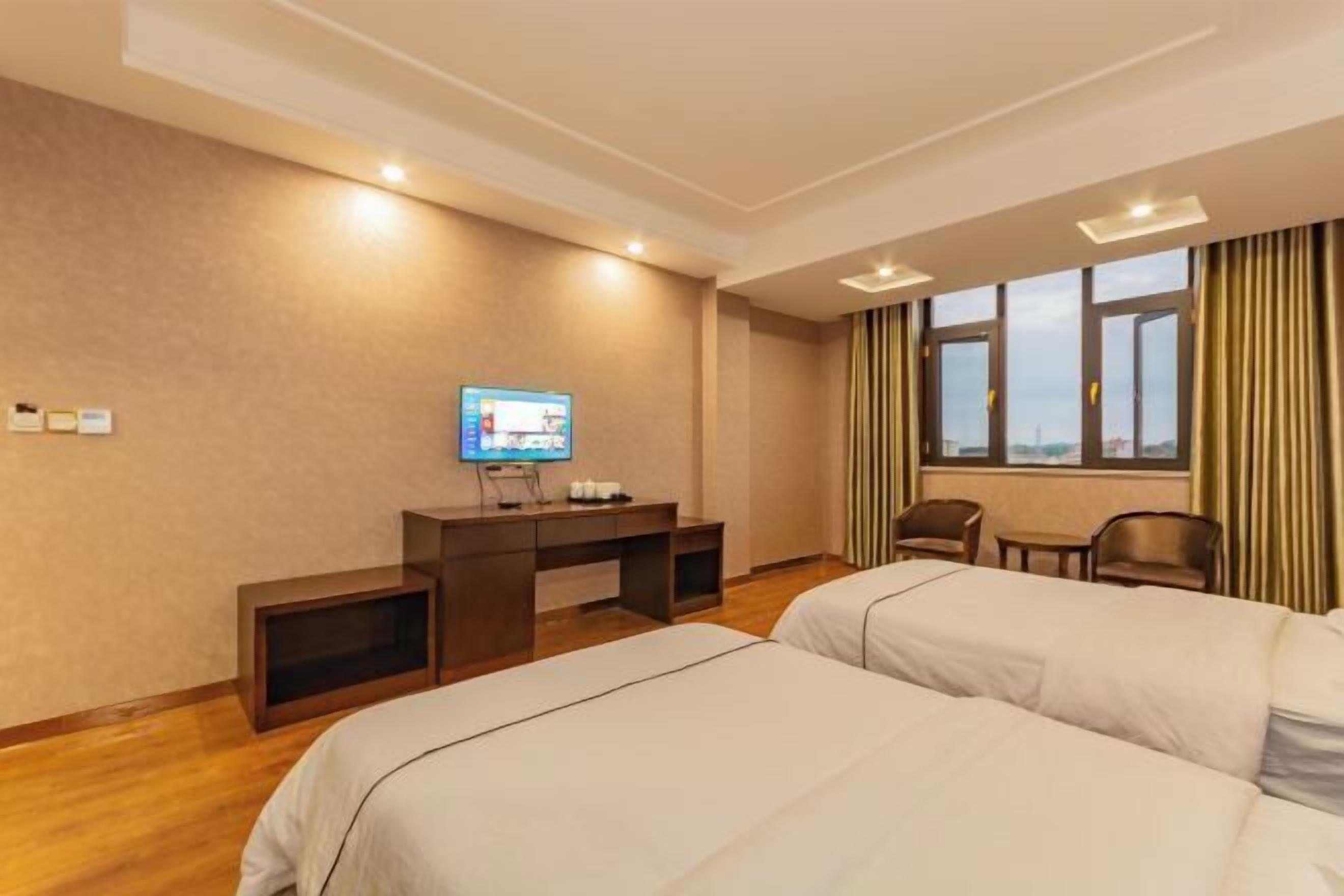 A hotel room with two beds, a desk with a television, a window with curtains, and a city view.