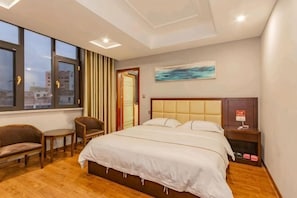 A hotel room with a large bed, a desk, a chair, a window with a city view, and a painting on the wall. - Fudu Hotel (Wuxue)