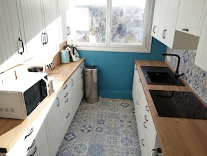 Una cocina compacta con armarios blancos, salpicadero de azulejos azules, ventana con vistas a edificios y un lavabo.