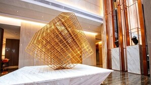 Um lobby moderno com uma escultura metálica geométrica sobre um pedestal de mármore, pisos de mármore polidos e um teto alto com iluminação embutida.