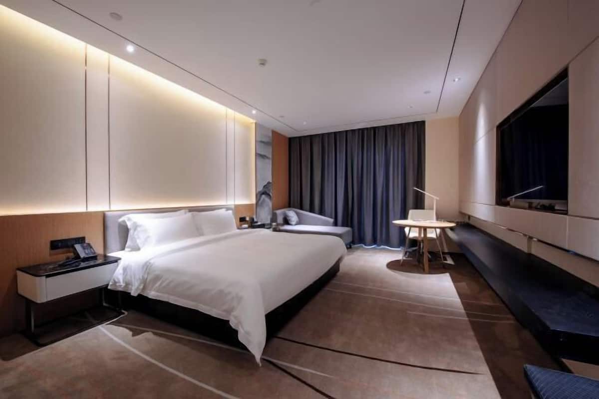 Ein modernes Hotelzimmer mit einem großen Bett, Nachttischen, einem Schreibtisch, einem Sessel, einem Fernseher und einem großen Fenster mit Vorhängen.