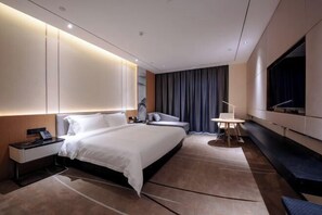A modern hotel room with a large bed, bedside tables, a desk, a chair, a TV, and a large window with curtains.