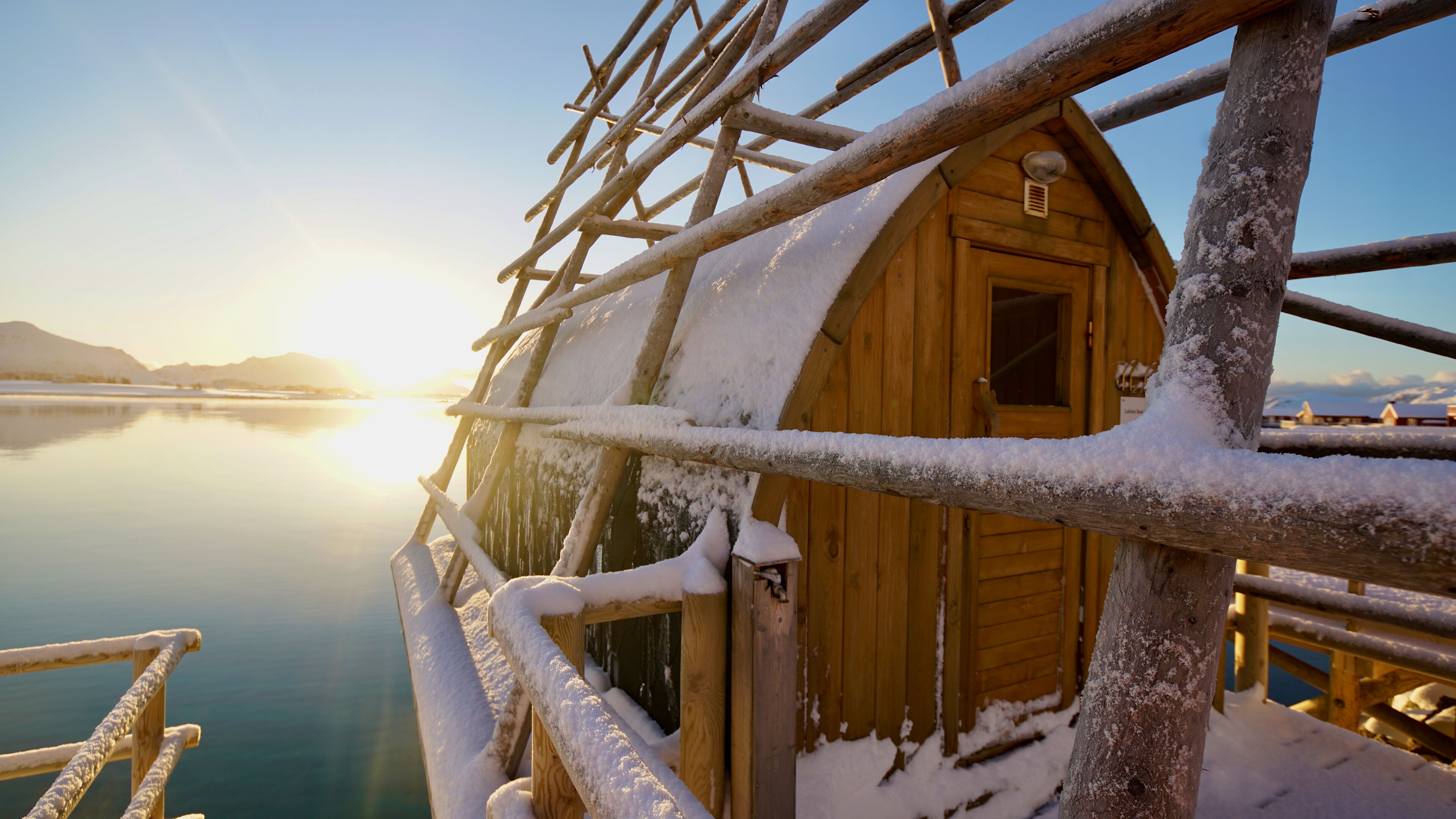 Une cabane en bois dont l’extérieur est recouvert de neige, située au bord d’une étendue d’eau calme, avec un ciel dégagé en arrière-plan.