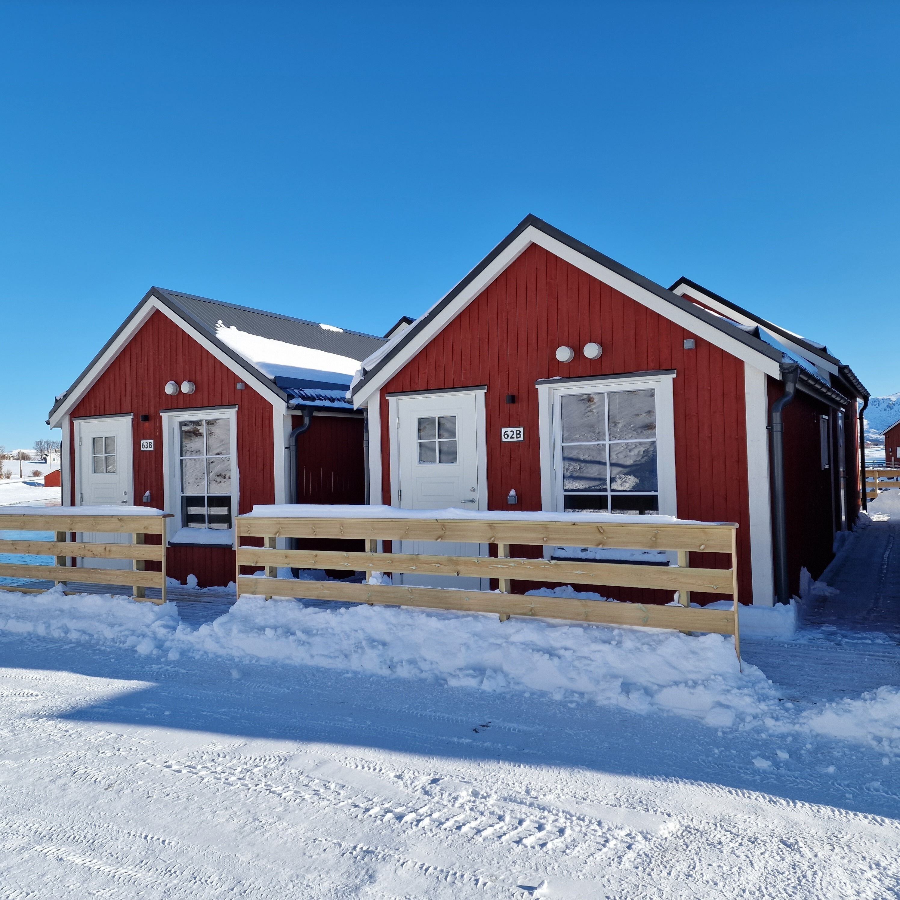 Un edificio rojo con dos puertas y ventanas, una cerca de madera en el frente, y nieve en el suelo.