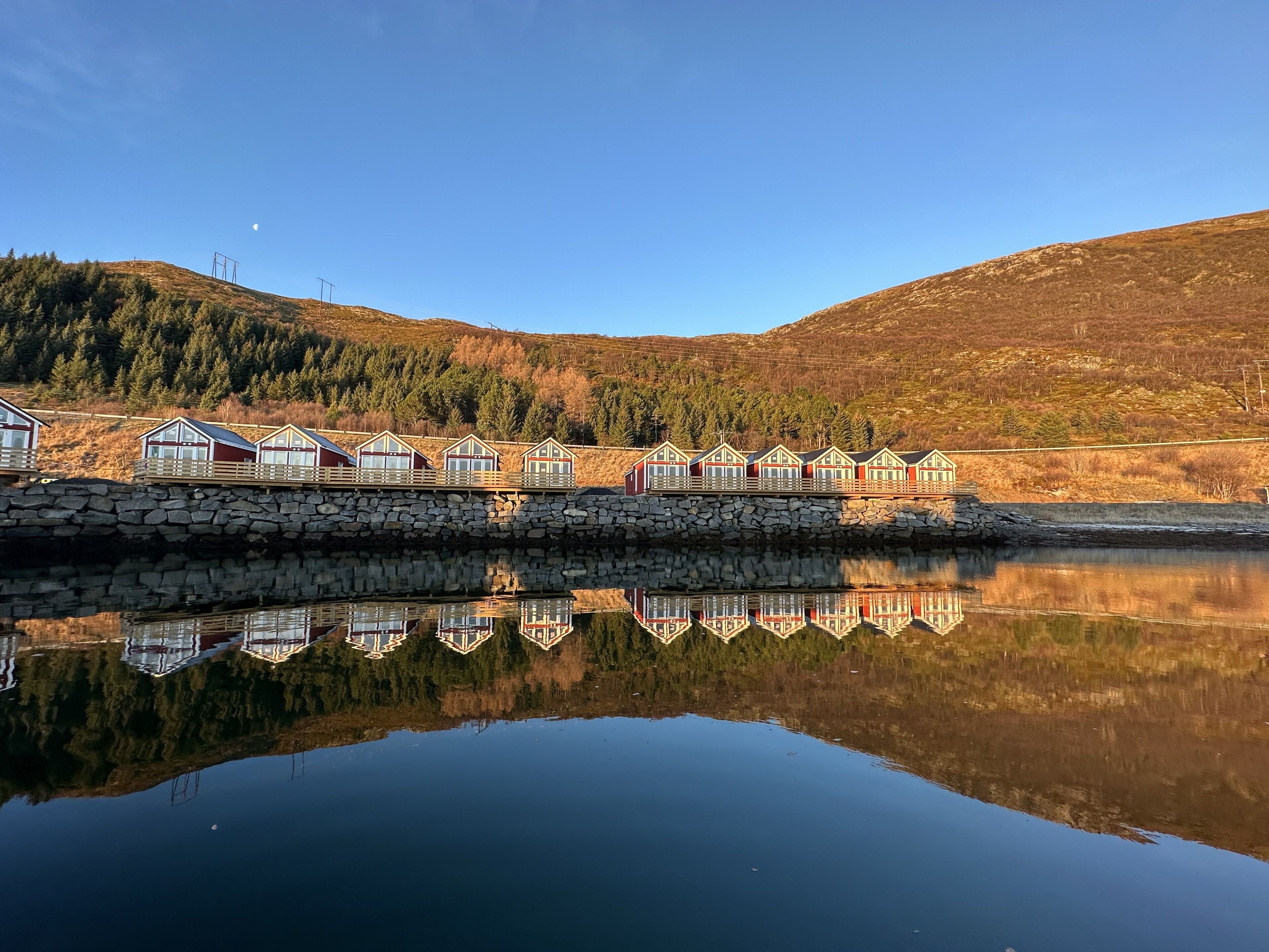 A row of cabins with a stone wall and a calm lake reflecting the structures.
