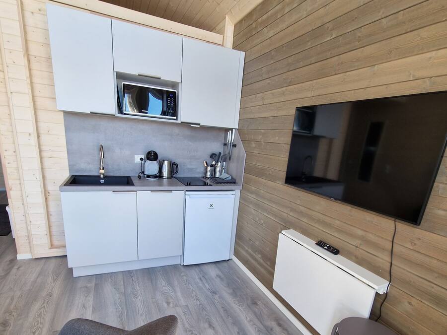 Cocina pequeña con armarios blancos, un microondas, una cafetera y un televisor montado en la pared. Hay dos taburetes y una silla en la zona de estar.