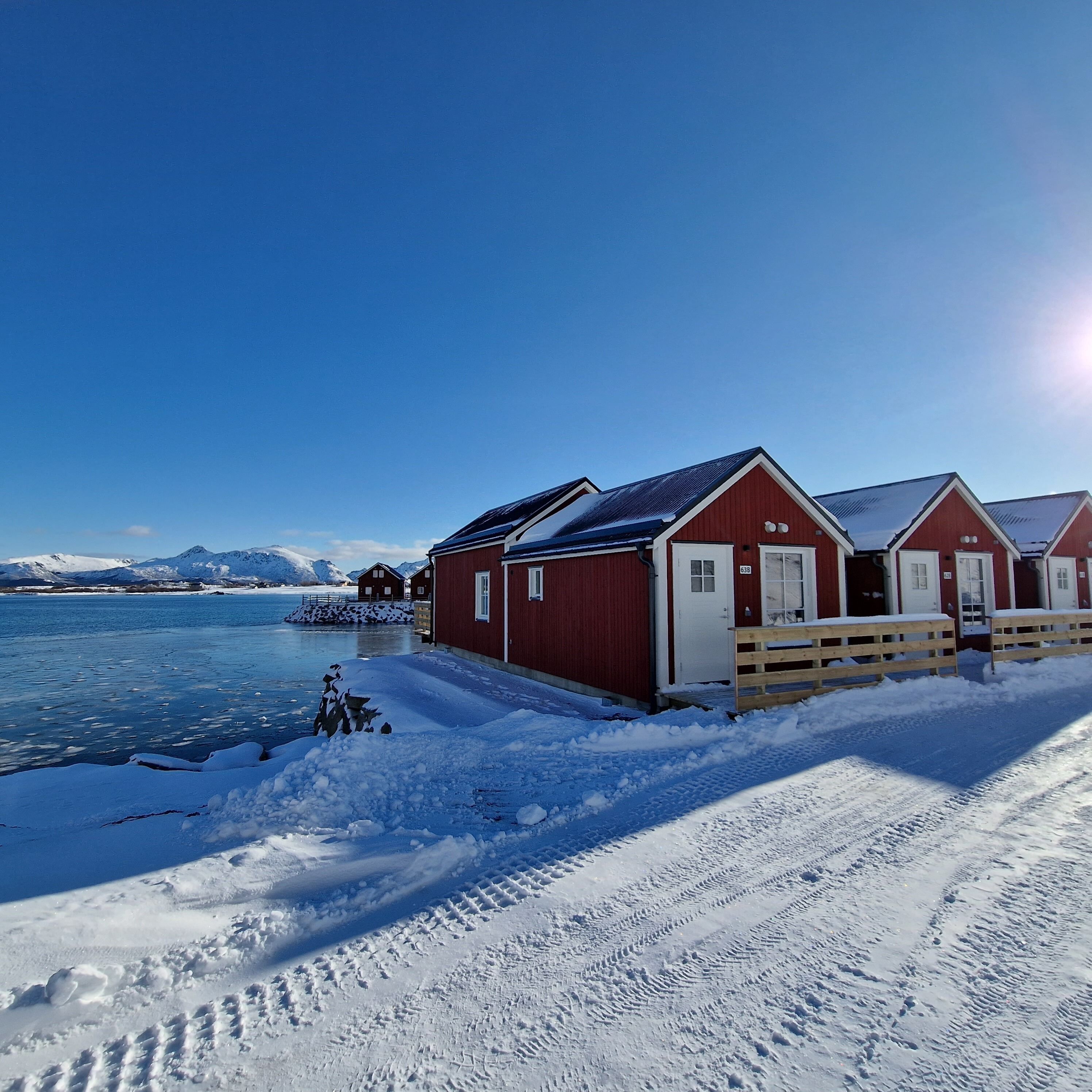 Une rangée de bâtiments en bois peints en rouge, avec des portes blanches, situés au bord d’un lac gelé, sous un ciel dégagé.