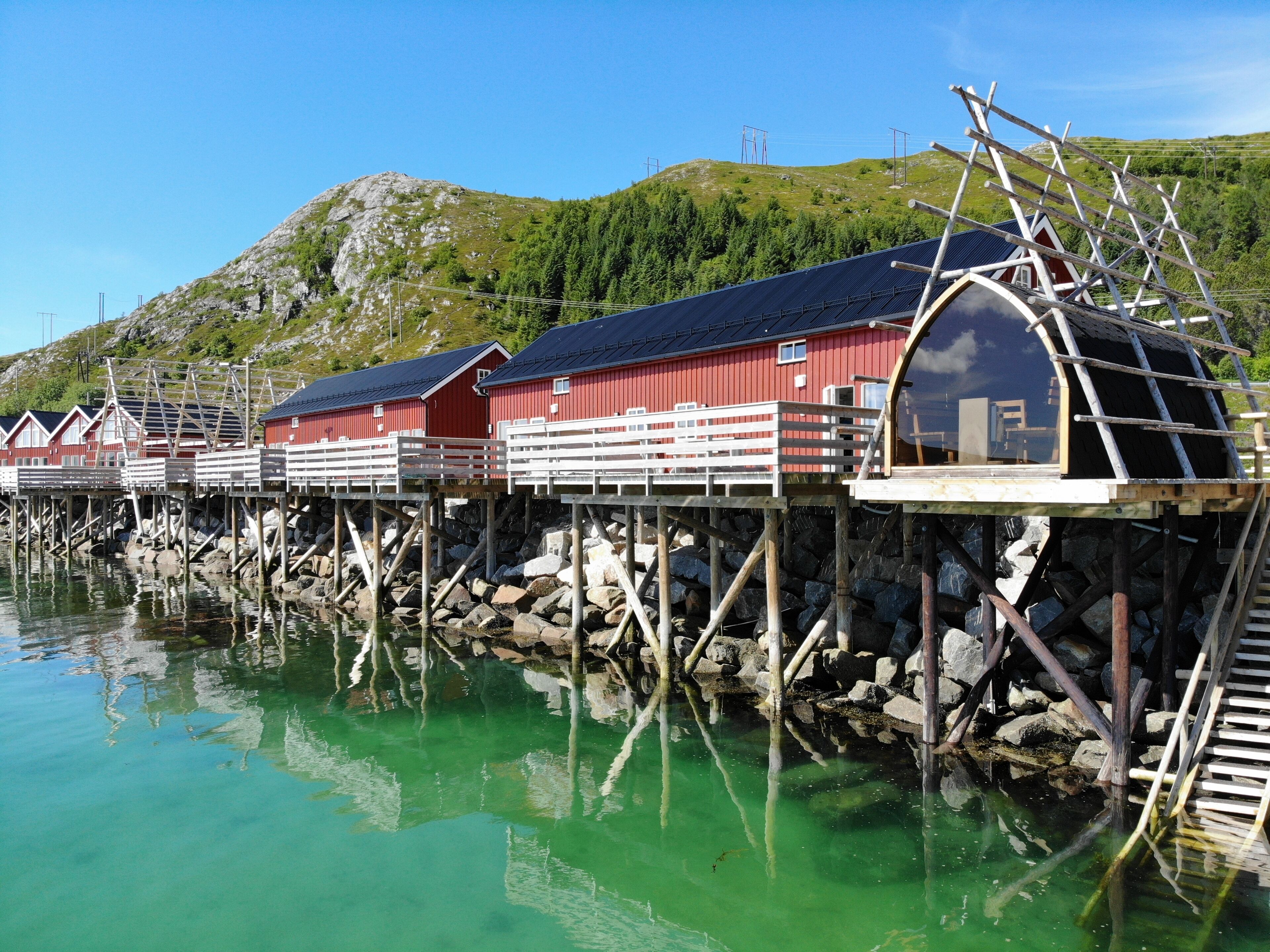 A row of red wooden buildings with white railings, connected by wooden walkways over water, with a mountainous backdrop.