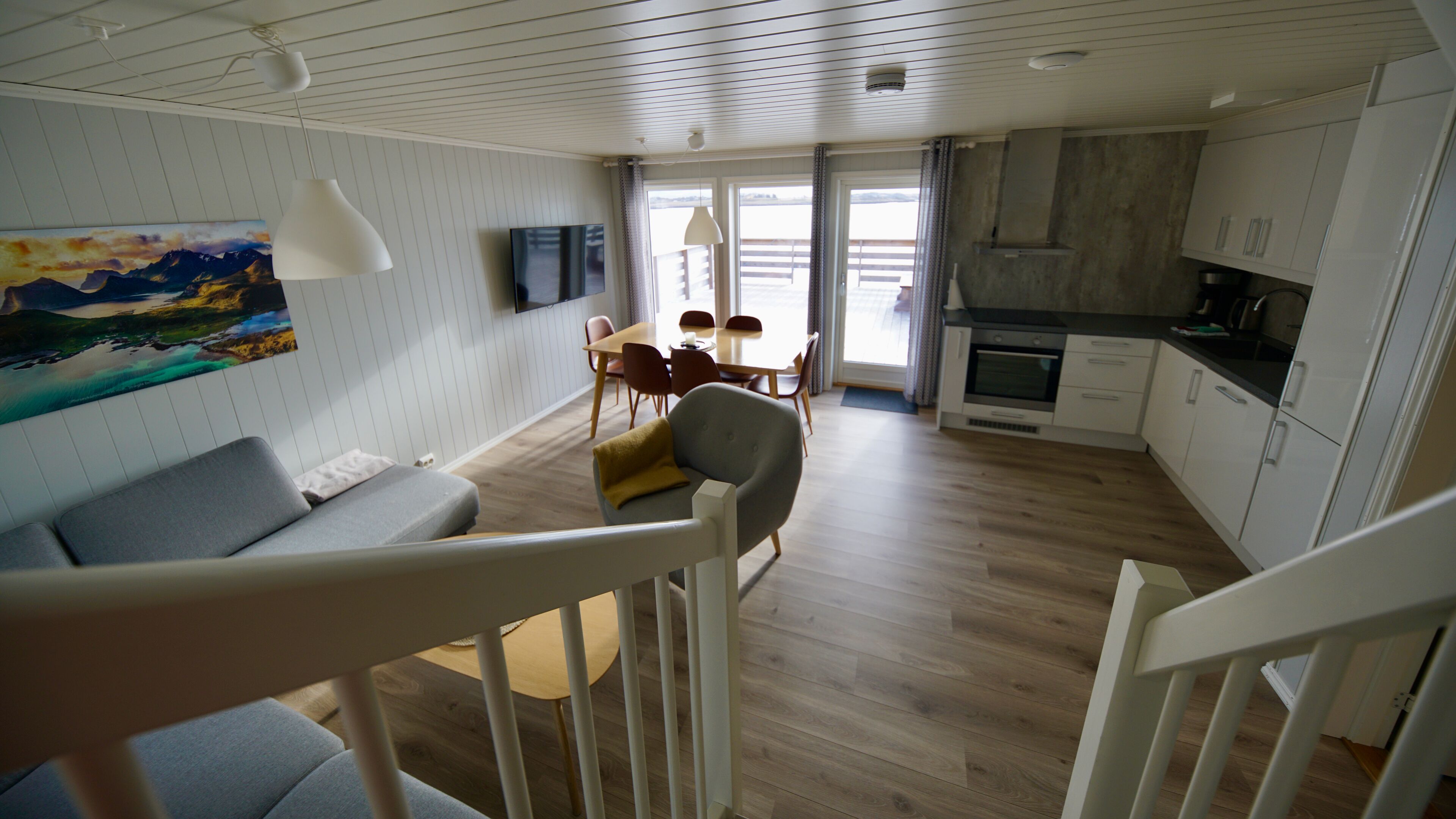A modern living room with a grey sofa, a dining table with chairs, a kitchen area with white cabinets, and a large window offering a view.