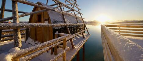 A wooden cabin with a snow-covered roof and a wooden deck extending over a body of water.