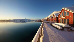 A row of red wooden buildings with snow-covered roofs, a calm lake, and a mountain in the background. - Lofoten Basecamp (Vestvågøy)
