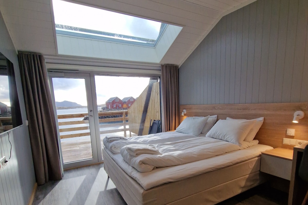 Ein Schlafzimmer mit Oberlicht, einem Bett, einem Nachttisch und Blick auf eine Holzterrasse sowie Gebäude.