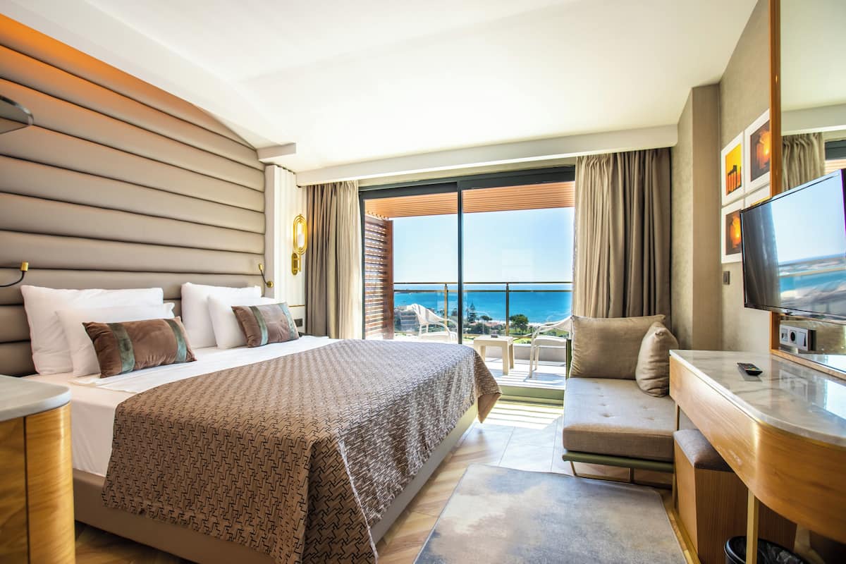 A hotel room with a large bed, a desk, a TV, and a balcony with a view of the ocean.