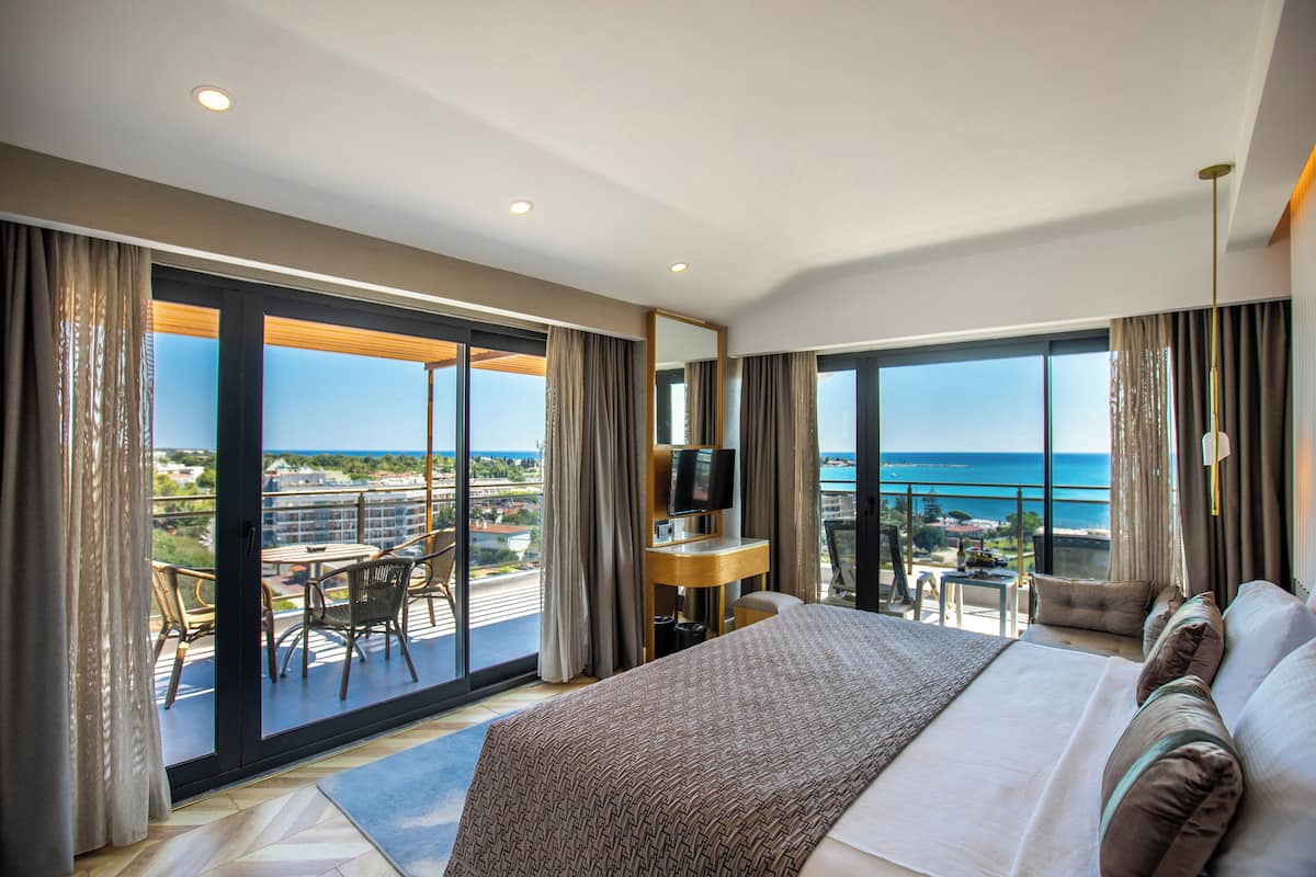 A hotel room with a large bed, a desk, and a TV. It has a balcony with a view of the ocean and buildings.