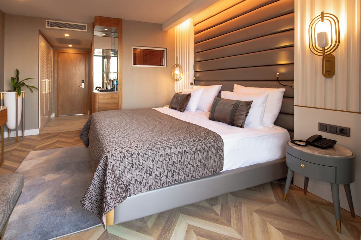 A modern hotel room with a large bed, a bedside table with a phone, a mirror, and a wall-mounted light fixture.