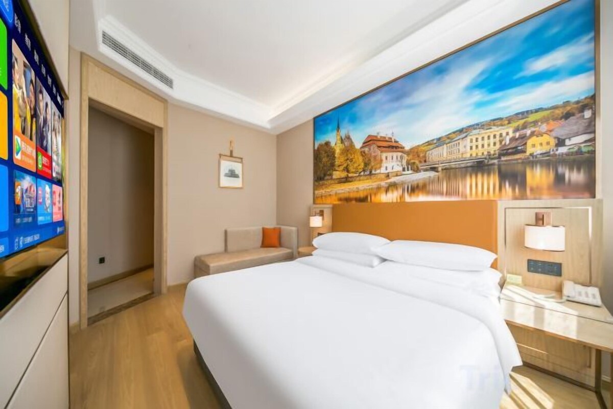 A hotel room with a large bed, a TV, a painting of a cityscape, and a desk with a lamp.