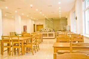 A spacious dining area with multiple wooden tables and chairs, a central buffet station, and a large window providing natural light. - Vienna Hotel (Zhoushan)