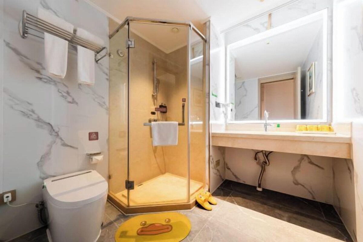 A bathroom with a glass-enclosed shower, a toilet, a sink with a mirror, and a yellow mat.
