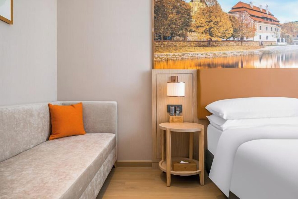 A hotel room with a bed, a sofa, a small table with a lamp, and a painting of a castle by a lake.