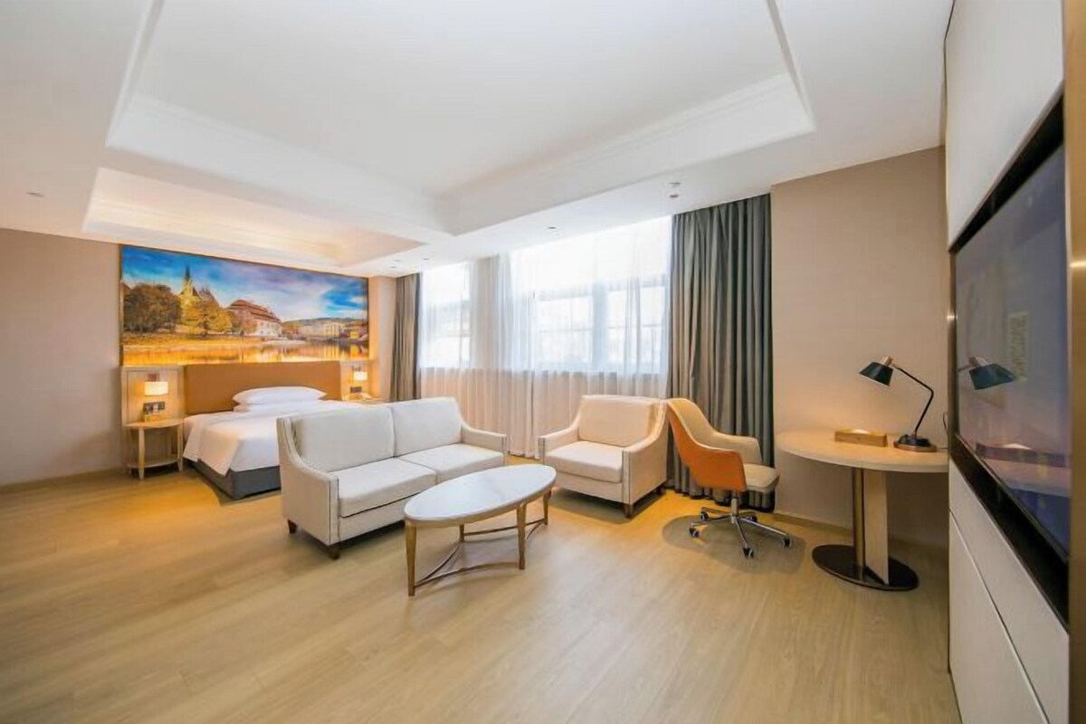 Une chambre d’hôtel moderne, dotée d’un grand lit, d’un canapé, d’une table basse et d’un bureau avec une chaise. Un tableau orne le mur et une fenêtre est agrémentée de rideaux.
