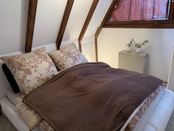 A bedroom with a bed, two patterned pillows, a brown blanket, a nightstand with a vase, and a window with a curtain. - Idyllic vacation home in Chiemgau (Siegsdorf)