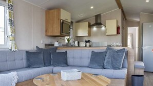 Un salon moderne avec un canapé d’angle gris, une table basse en bois et un coin cuisine doté de meubles en bois et d’un four à micro-ondes.