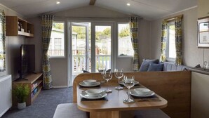 Un coin repas accueillant avec une table en bois dressée pour quatre personnes, de grandes fenêtres avec des rideaux, un téléviseur posé sur une étagère et une vue sur l’extérieur.