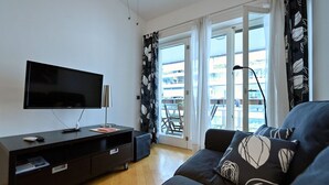 Una sala moderna con un televisor de pantalla plana, un sofá de color oscuro con cojines estampados, piso de madera y un cerramiento de vidrio corredizo que da a un balcón exterior.