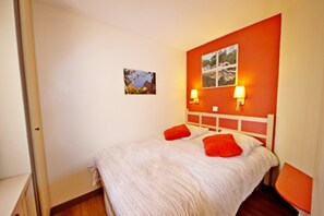 Quarto com cama, duas almofadas vermelhas, cabeceira branca, mesa de cabeceira e duas luminárias fixadas na parede.