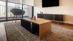 A modern living room with a wooden coffee table, a black armchair, a TV mounted on the wall, and a large window with a view of the outside.