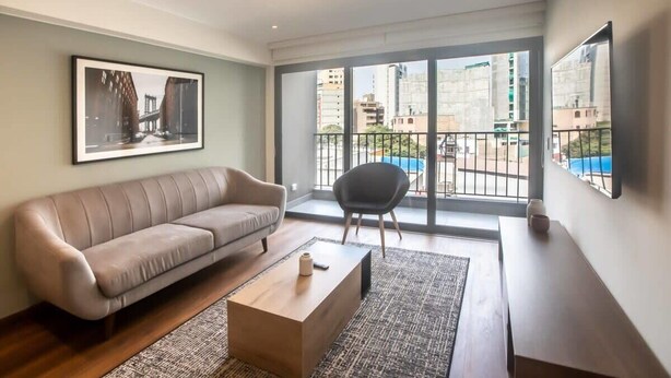 A modern living room with a sofa, a coffee table, and a chair. There is a large window with a view of buildings and a balcony.
