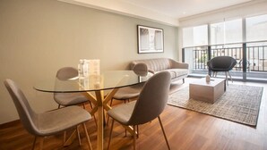 A modern living room with a glass dining table, grey upholstered chairs, a grey sofa, and a black chair. There is a large window with a balcony.