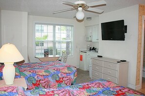 A bedroom with a bed, a desk, a chair, a lamp, a ceiling fan, a TV, and a window with blinds.