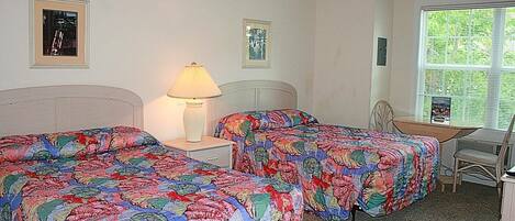 A room with two beds, a desk, and a chair. There is a ceiling fan, a lamp, and a window with blinds.