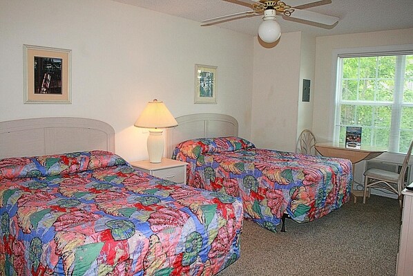 A room with two beds, a desk, and a chair. There is a ceiling fan, a lamp, and a window with blinds.