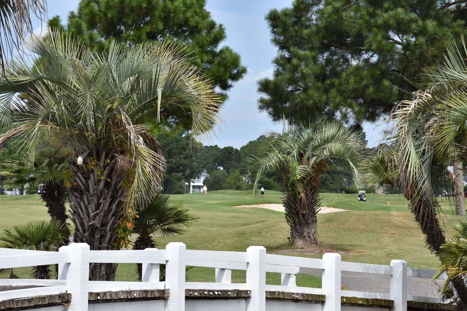 A golf course with a white fence, palm trees, and a clubhouse in the distance.