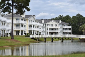 A multi-story hotel with balconies, a dock over water, and a grassy area. - Brunswick Plantation Resort and Golf Villa 2705L in the Heart of NC Seafood Country by RedAwning (Calabash)