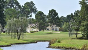 A golf course with a pond, trees, and a house in the background. - 2609l Home by RedAwning (Calabash)