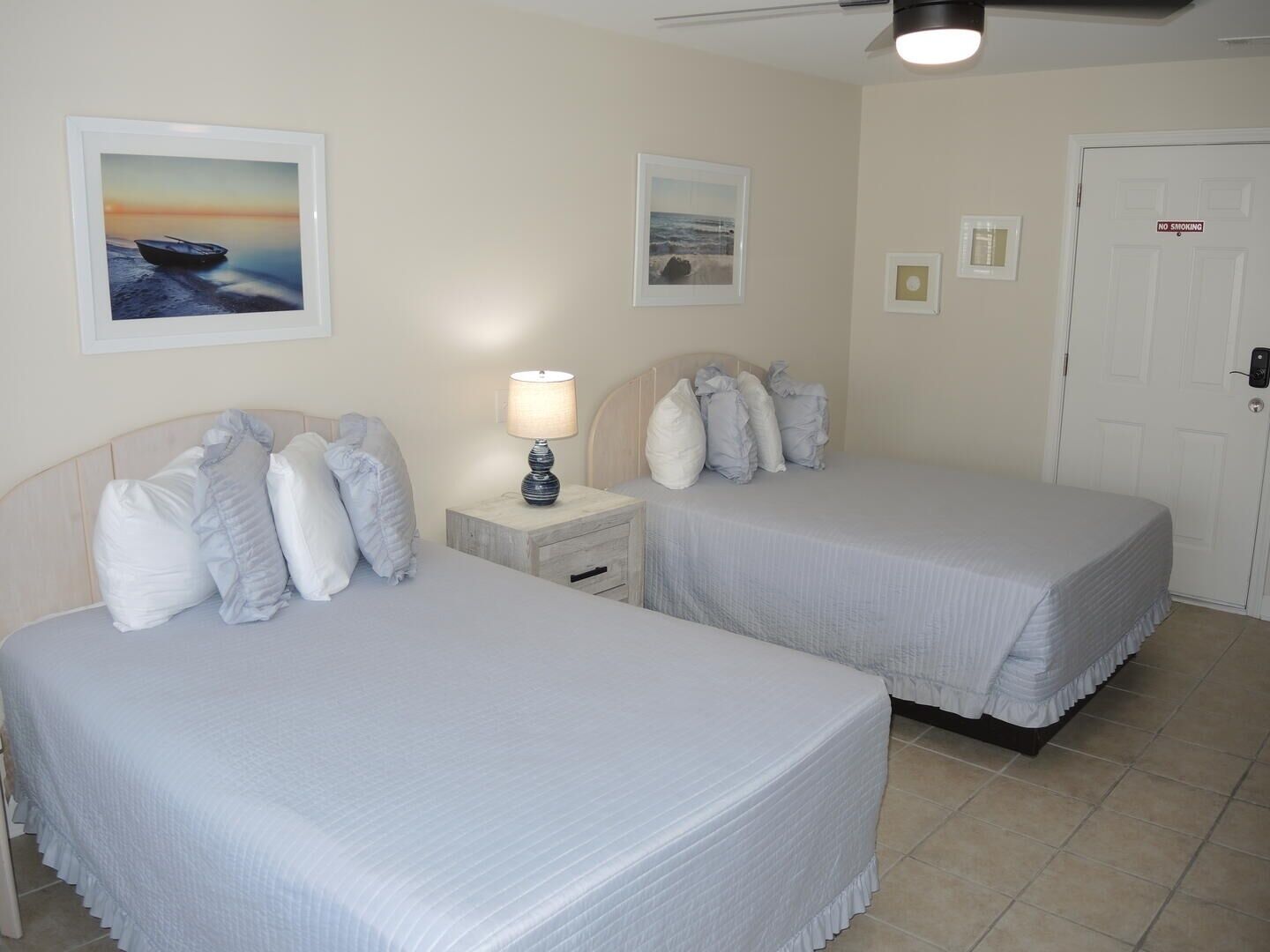 A hotel room with two beds, a nightstand with a lamp, and a door with a 'No Smoking' sign.