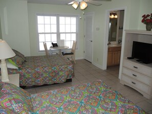 A bedroom with two beds, a desk, a chair, a television, and a bathroom visible through an open door.