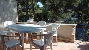 Een terras buiten met een ronde tafel en witte plastic stoelen.