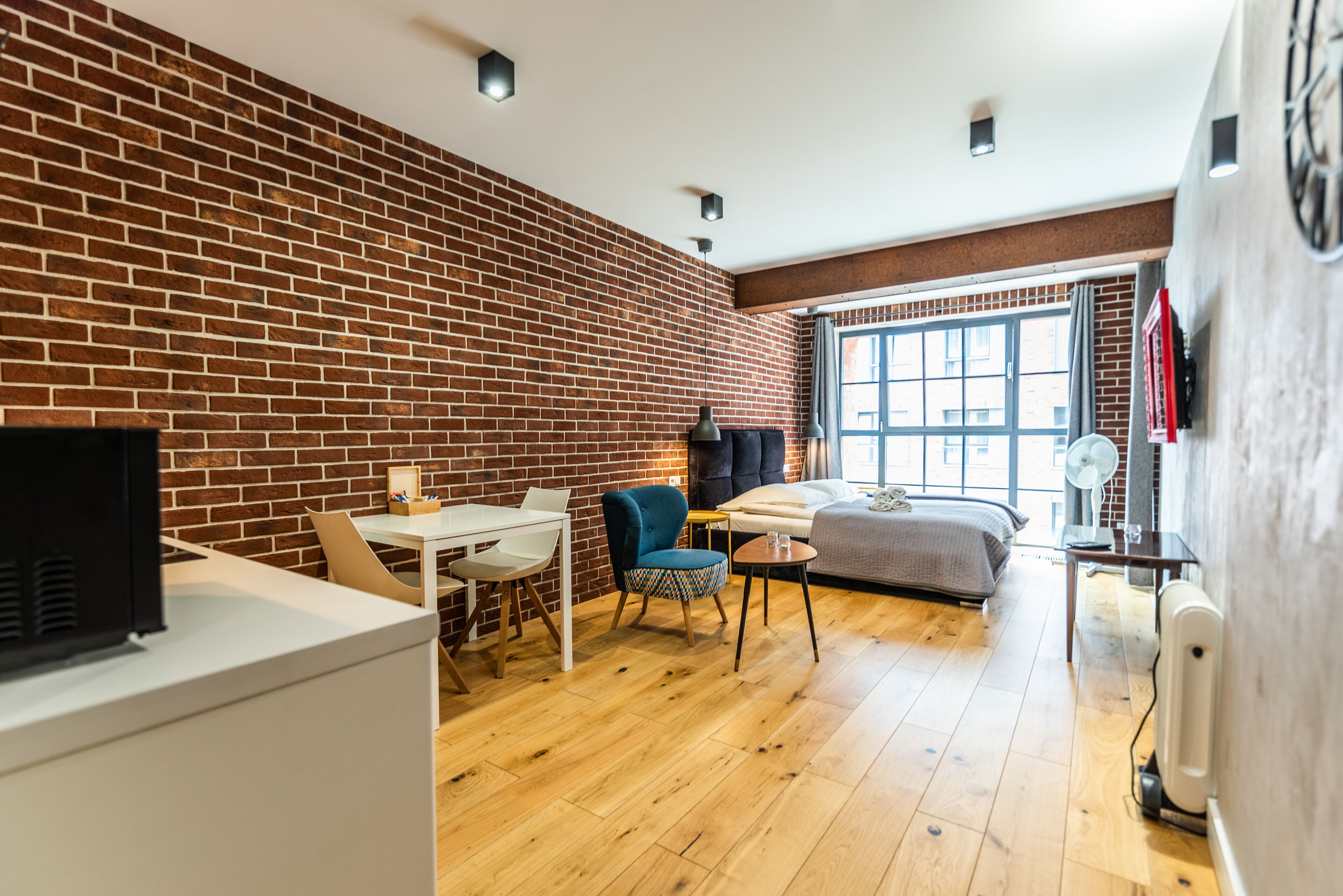 Una habitación moderna con pared de ladrillo, suelo de madera, cama, escritorio con silla, una mesita y ventana con cortinas.