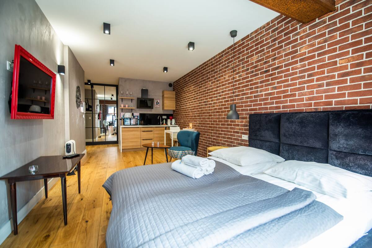Une chambre d’hôtel moderne avec un mur en briques, un grand lit recouvert d’une couette grise, un bureau en bois et, au fond, un coin cuisine.